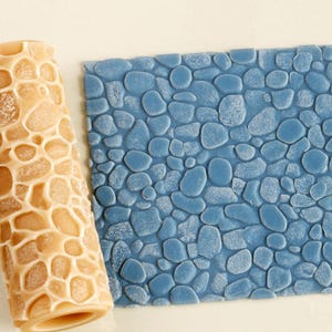 May include: A cylindrical tan rolling pin with a raised, irregular grid pattern designed for texturing, positioned next to a square piece of blue clay that displays a clear, embossed impression of rounded river stones or pebbles.