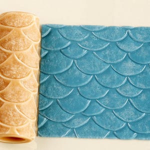 Op de afbeelding: Een cilindrische, beige deegroller met een verhoogd vissenschubpatroon naast een vierkant stuk blauw materiaal dat is voorzien van hetzelfde herhalende schubontwerp, wat de textuur laat zien die door het gereedschap is aangebracht.