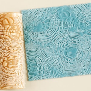 Puede incluir: Un rodillo cilíndrico de color tostado con patrón, situado junto a una pieza cuadrada de material azul claro. El rodillo presenta una textura intrincada en relieve que crea un efecto de ondas, impreso claramente en la superficie azul.