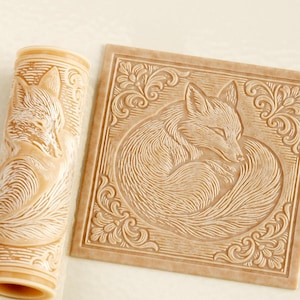 May include: A light tan cylindrical rolling pin and a matching square flat stamp, both featuring a deeply embossed design of a curled fox surrounded by intricate floral scrollwork patterns, rendered in a monochromatic light brown tone.