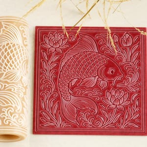 May include: A cylindrical beige rolling pin featuring an embossed koi fish and lotus flower pattern, positioned next to a square red tile displaying the same intricate relief design of a swimming koi fish surrounded by blooming lotus flowers.