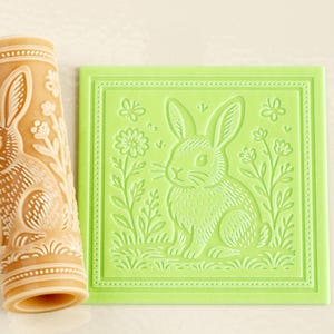 May include: A tan cylindrical rolling pin and a square light green flat stamp, both featuring an embossed design of a rabbit sitting among flowers and butterflies. The rolling pin has a repeating pattern, while the stamp shows a single centered rabbit.