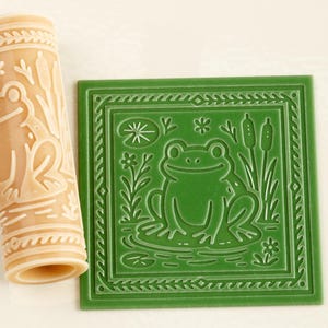 May include: A cylindrical beige rolling pin with a raised frog and cattail pattern alongside a square green tile featuring an identical embossed frog, cattail, and lily pad design. The tools are designed for creating detailed impressions in clay.