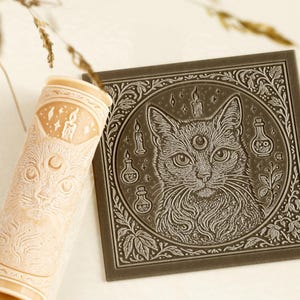 May include: A cylindrical beige rolling pin and a square dark gray tile, both featuring an embossed design of a cat with a third eye on its forehead, surrounded by candles, small bottles, and botanical elements within a circular frame.