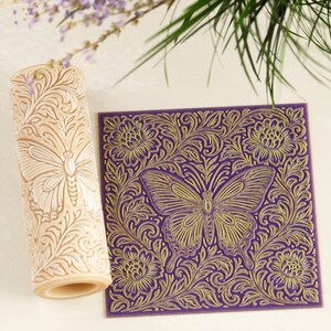 May include: A beige rolling pin and a purple square with a butterfly and floral design. The rolling pin has a butterfly and floral pattern. The square has a gold butterfly and floral design on a purple background.