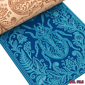May include: A light brown rolling pin with an embossed floral design rests on a blue mat with a detailed beetle and botanical pattern. The mat features a square shape with a border, and the text "STL FILE" is visible in the corner.