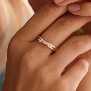 Peut inclure: Une bague croisée en or ornée de cinq petites pierres précieuses rondes et multicolores alignées au centre. L'anneau présente une finition polie et un design délicat et entrelacé qui crée un aspect superposé sur le doigt.