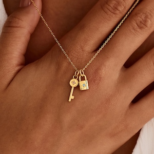 May include: A delicate gold necklace featuring a key and a padlock pendant. The key has a circular head, and the padlock has a small gemstone. The necklace is displayed on a person's chest, with the chain visible.