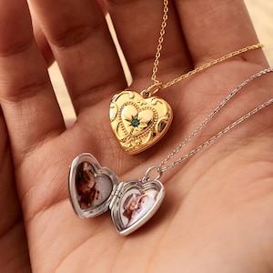 May include: A gold heart-shaped locket with an engraved pattern and a small central teal stone on a thin chain, alongside an open silver heart-shaped locket on a thin chain containing two small photographs inside the metal frame.