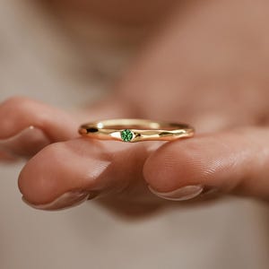 Peut inclure: Une bague en or avec une petite pierre verte ronde au centre. La bague est tenue dans une main, mettant en valeur son design et sa couleur. Un bijou délicat, idéal pour un cadeau ou un usage quotidien.