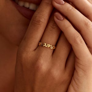 May include: A gold-toned ring featuring the word mama in a cursive, script-style font. The ring is thin and polished, with the letters connected to form the band. The design is simple and metallic, showcasing a smooth, reflective surface finish.