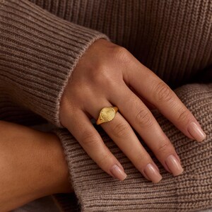 K&ouml;nnte beinhalten: Ein goldener Siegelring mit strukturiertem, kreisf&ouml;rmigem Design. Der Ring wird an einer Hand mit manik&uuml;rten N&auml;geln getragen, vor einem braunen Pullover.