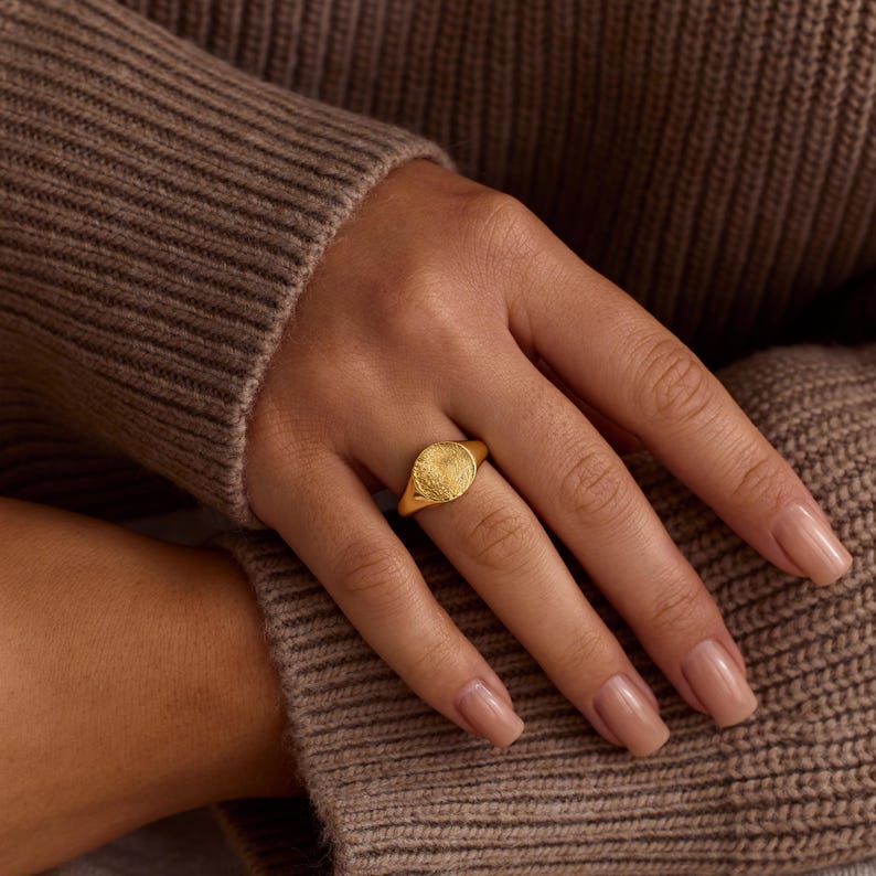 K&ouml;nnte beinhalten: Ein goldener Siegelring mit strukturiertem, kreisf&ouml;rmigem Design. Der Ring wird an einer Hand mit manik&uuml;rten N&auml;geln getragen, vor einem braunen Pullover.