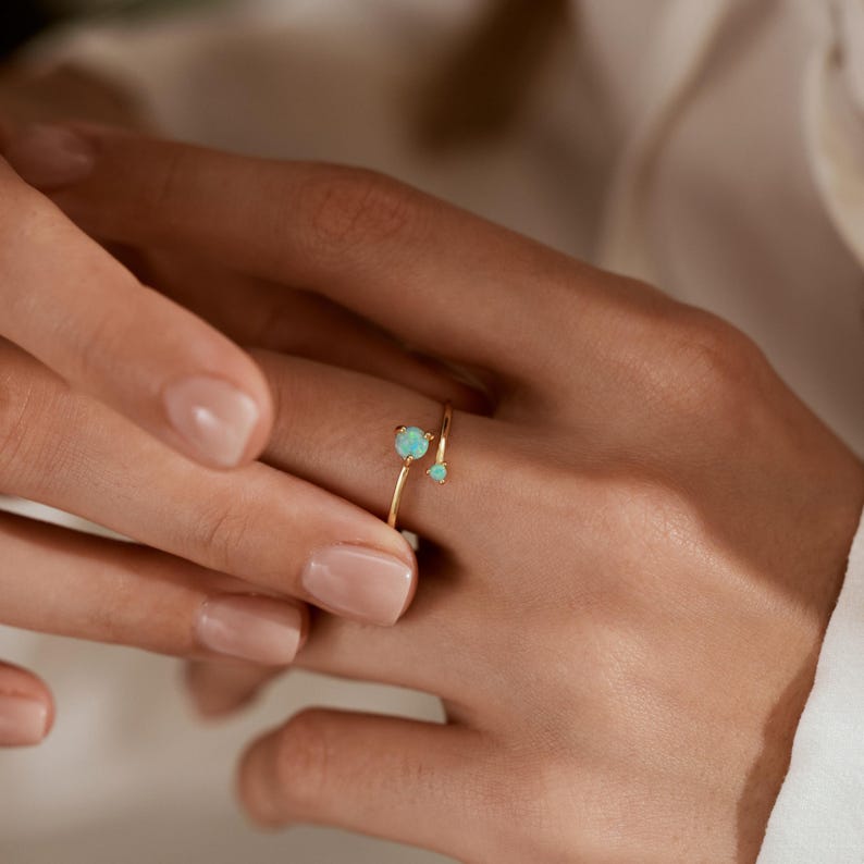 May include: A delicate gold ring featuring two light blue gemstones. The ring has a minimalist design with a slender band that wraps around the finger. The gemstones are round and set in a simple, elegant style.