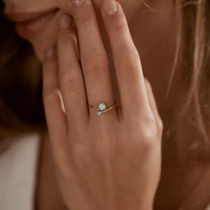 May include: A gold-colored ring with two small, light blue gemstones. The ring has a delicate, minimalist design, with the gemstones set on a thin band. The ring is shown on a person's finger.