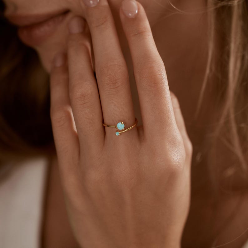May include: A gold-colored ring with two small, light blue gemstones. The ring has a delicate, minimalist design, with the gemstones set on a thin band. The ring is shown on a person's finger.