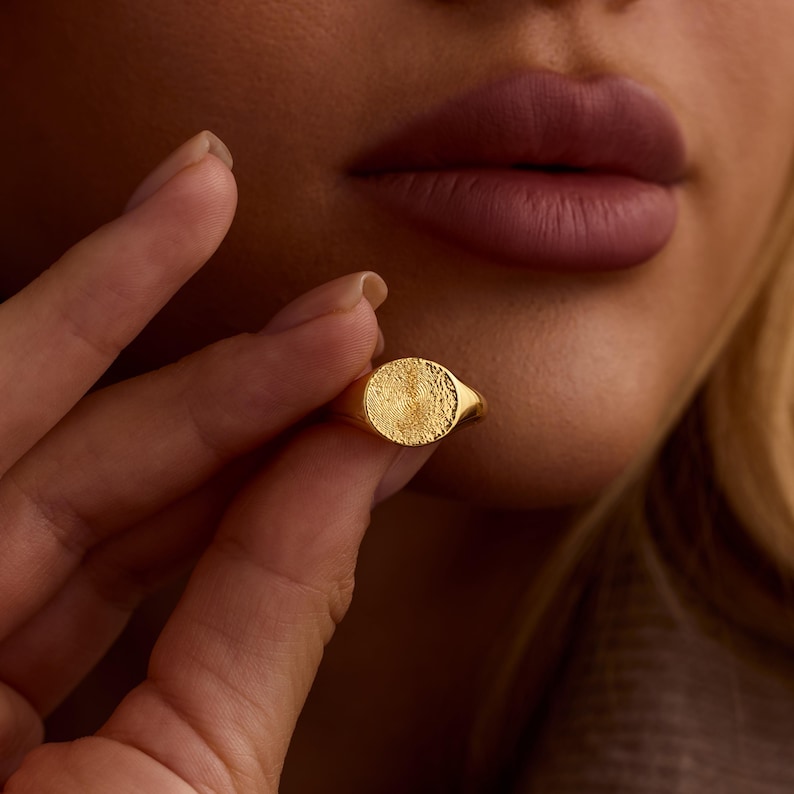 K&ouml;nnte beinhalten: Ein goldener Siegelring mit einer strukturierten, kreisf&ouml;rmigen Oberfl&auml;che. Der Ring wird von den Fingern einer Person gehalten, mit einem verschwommenen Hintergrund von Gesicht und Kleidung einer Person. Der Ring ist ein klassisches Schmuckst&uuml;ck.