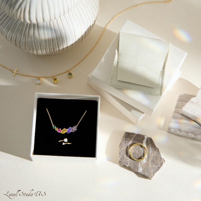 K&ouml;nnte beinhalten: Eine Sammlung von Goldschmuck, darunter Halsketten und Ringe, wird auf einer wei&szlig;en Oberfl&auml;che pr&auml;sentiert. Eine Halskette hat einen Kreuzanh&auml;nger, w&auml;hrend eine andere farbige Edelsteine aufweist. Ein goldener Ring liegt auf einem Stein.