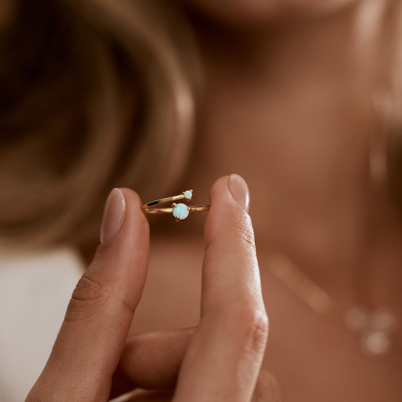 May include: A gold-colored ring with two light blue opal stones. The ring has a delicate, open design, with one stone set on the main band and a smaller stone on a separate, curving band. The ring is held up by a person's fingers.