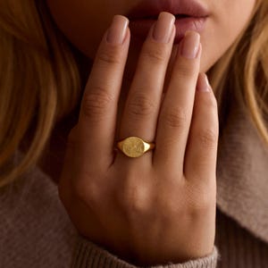 K&ouml;nnte beinhalten: Ein goldener Siegelring mit einer strukturierten, kreisf&ouml;rmigen Oberfl&auml;che. Der Ring wird an einem Finger getragen, wobei die Hand und ein Teil des Gesichts sichtbar sind. Das Design des Rings ist schlicht und elegant, f&uuml;r den Alltag geeignet.