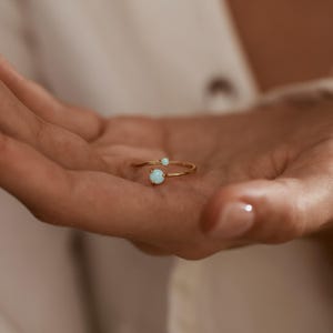 May include: A gold-colored ring with two light blue opal stones. The ring has a delicate, open design, with one opal at the end of the band and a smaller one further up. The ring is shown on a person's hand.