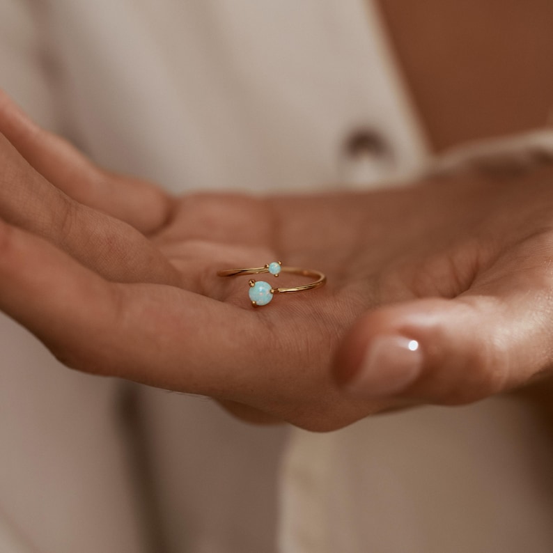 May include: A gold-colored ring with two light blue opal stones. The ring has a delicate, open design, with one opal at the end of the band and a smaller one further up. The ring is shown on a person's hand.