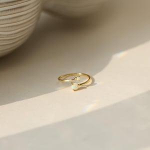 May include: A gold-colored ring with two small, oval-shaped, white gemstones. The ring has a delicate, open design, with one gemstone at the end of each band. The ring is set against a neutral background, with soft lighting.