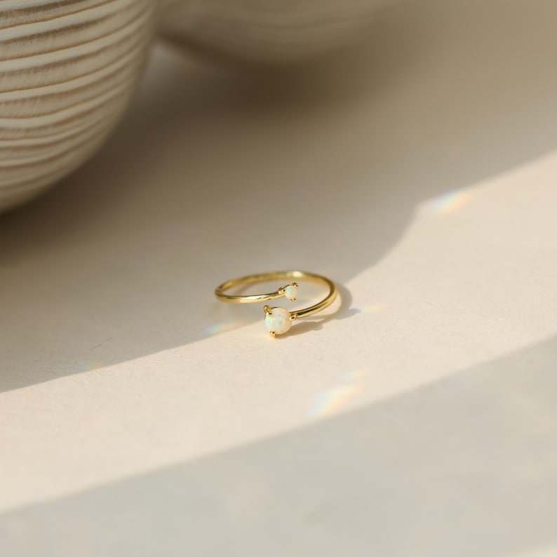 May include: A gold-colored ring with two small, oval-shaped, white gemstones. The ring has a delicate, open design, with one gemstone at the end of each band. The ring is set against a neutral background, with soft lighting.