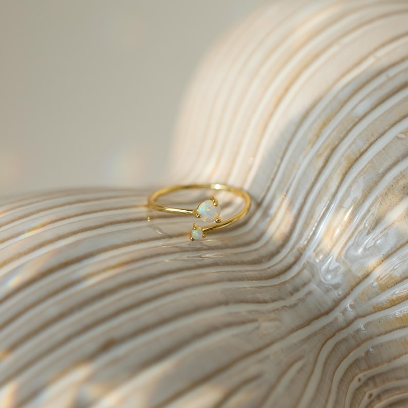 May include: A delicate gold ring with two opal stones. The ring features a slender band that curves around the finger, with a larger opal stone at the top and a smaller one below. The opals have a milky white color.