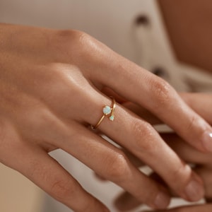 May include: A delicate gold ring featuring two opal gemstones. The ring has a slender, curved design with a larger opal at the top and a smaller one below. The opals have a milky white color with subtle iridescent flashes.