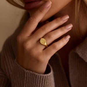 K&ouml;nnte beinhalten: Ein goldener Siegelring mit einer strukturierten ovalen Oberfl&auml;che. Der Ring wird an einem Finger getragen, wobei die Hand auf einem neutralfarbenen Pullover ruht. Das Design des Rings ist schlicht und elegant.