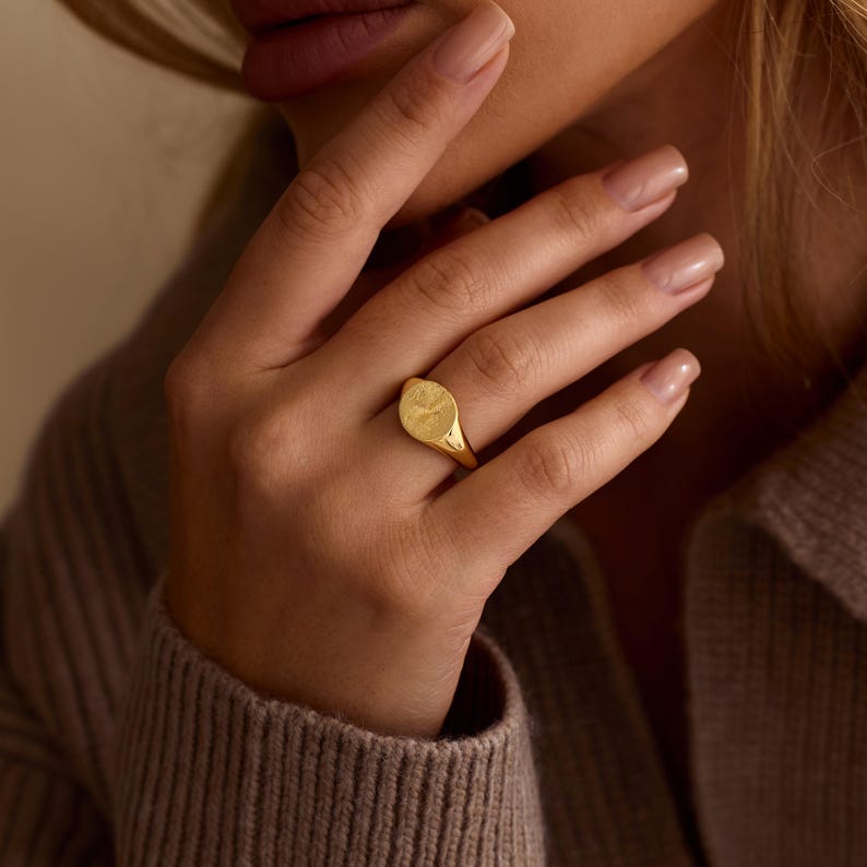 K&ouml;nnte beinhalten: Ein goldener Siegelring mit einer strukturierten ovalen Oberfl&auml;che. Der Ring wird an einem Finger getragen, wobei die Hand auf einem neutralfarbenen Pullover ruht. Das Design des Rings ist schlicht und elegant.