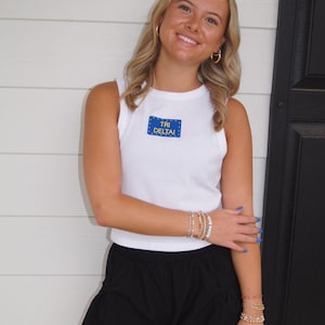 May include: A woman wearing a white tank top with a blue and yellow "TRI DELTA" patch, paired with black shorts. She is also wearing gold hoop earrings and multiple bracelets. The background features a white wall and a black door.