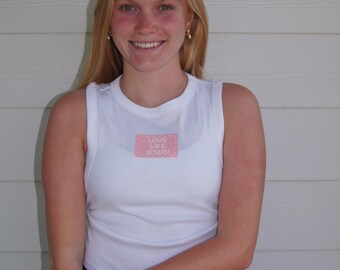 May include: A white tank top with the text "LOVE LIKE JESUS!" on a pink square. The person is wearing a black skirt and a gold bracelet. The background is a white wooden wall.