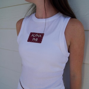 May include: White sleeveless top with the text "ALPHA PHI" on a maroon patch. The top is paired with white shorts. The image is taken in natural light.