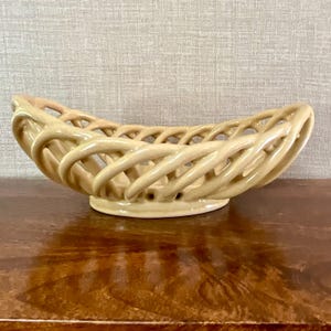May include: A beige ceramic bowl featuring an openwork lattice design. The bowl has a rounded, elongated shape with a smooth, glossy finish and a solid base, constructed from interconnected, curved ceramic strands that form a basket-like structure.