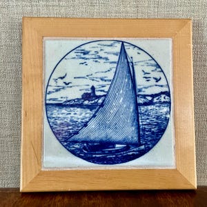 May include: A square, framed tile featuring a blue and white nautical scene. The image depicts a sailboat with a large sail, a lighthouse, and birds flying in the sky. The tile is set in a light-colored wooden frame.