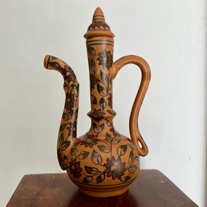 May include: A tall, light brown ceramic pitcher with a long, slender neck, a curved handle, and a pointed lid. The surface is decorated with hand-painted dark brown and green floral patterns featuring blooming flowers and leafy vines throughout.