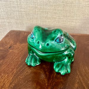 May include: A ceramic frog-shaped trinket box. The frog is a glossy, vibrant green with blue eyes. The box is detailed with textured skin and a hinged lid, perfect for storing small items. The frog is sitting on a wooden surface.