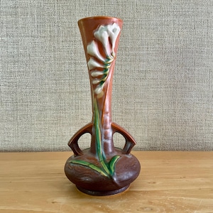 May include: A tall, brown ceramic vase with a flared rim and two side handles. The vase features a raised, white floral relief design extending from the base up the neck, accented with green stems and leaves against a smooth, glazed brown surface.