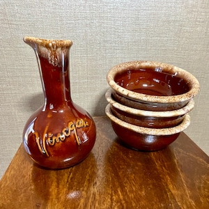 May include: A brown ceramic vase with a curved spout and the word "Vinegar" in cursive script. Beside it, a stack of three brown ceramic bowls with a lighter rim.