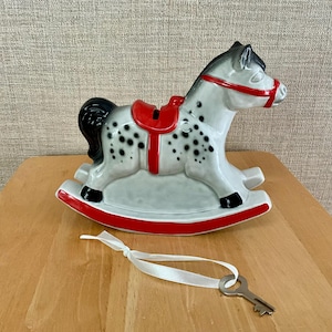 May include: A ceramic rocking horse piggy bank featuring a light gray body with black spots, a black mane and tail, and a bright red saddle, bridle, and rocker base. A white ribbon is tied to the horse, attached to a small silver-colored metal key.