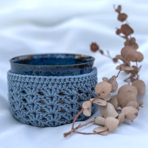 Könnte beinhalten: Eine blaue Keramiktasse mit strukturiertem, dunkelblauem Innenraum sitzt in einem hellblauen gehäkelten Becherhalter. Der Becherhalter hat ein offenes, gemustertes Design. Getrocknete braune Blätter und Stiele sind im Hintergrund.