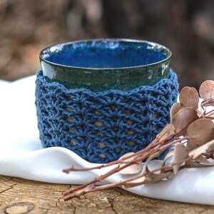Könnte beinhalten: Eine handgefertigte Keramiktasse mit blauer Glasur, umhüllt von einer gehäkelten Hülle. Die Hülle ist tiefblau und hat ein strukturiertes Stichmuster. Die Tasse und die Hülle stehen auf einem weißen Tuch, mit getrockneten Zweigen im Vordergrund.
