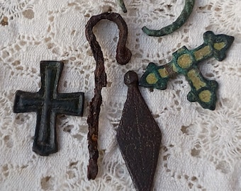 May include: A collection of five metal artefacts including a dark cross, a rusted shepherd's crook, a diamond-shaped pendant with etched patterns, a green oxidised ring, and a decorative cross with yellow-toned accents on a white lace background.