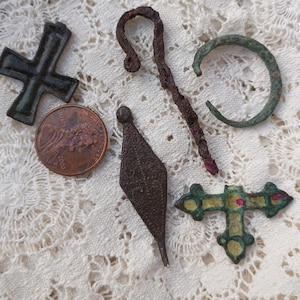 May include: A collection of five metal artefacts including a dark cross, a rusted shepherd's crook, a green circular band, a diamond-shaped pendant, and a decorative cross with red accents, displayed alongside a copper penny for scale.