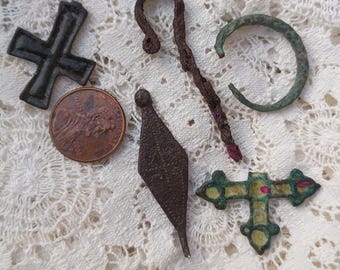Op de afbeelding: Een verzameling van vijf metalen voorwerpen, waaronder een donker kruis, een roestige herdersstaf, een groene cirkelvormige ring, een ruitvormige hanger en een decoratief kruis met rode accenten, getoond naast een koperen munt.