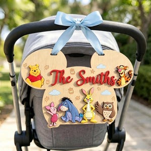 May include: A light wood sign shaped like a mouse head, featuring Winnie the Pooh, Tigger, Piglet, Eeyore, Rabbit, and Owl. The sign displays the red text "The Smiths" and is decorated with small honey pots, leaves, and clouds, topped with a light blue ribbon.