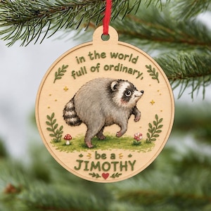 Puede incluir: Un adorno de madera redondo con una ilustraci&oacute;n impresa de un mapache sobre hierba verde. El texto dice: "in the world full of ordinary, be a JIMOTHY". El dise&ntilde;o incluye peque&ntilde;as ramas verdes, una seta roja y una peque&ntilde;a flor.