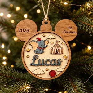 May include: A wooden Mickey-shaped Christmas ornament featuring a layered design with the name Lucas, a Dumbo elephant illustration, a circus tent, clouds, stars, a red ball, the year 2026, and the text 1st Christmas, suspended by a twine loop.